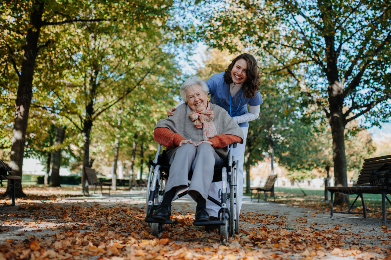 Disabled elderly woman in wheelchair on walk in park during warm autumn day. Young nurse pushing wheelchair, talking with senior woman.