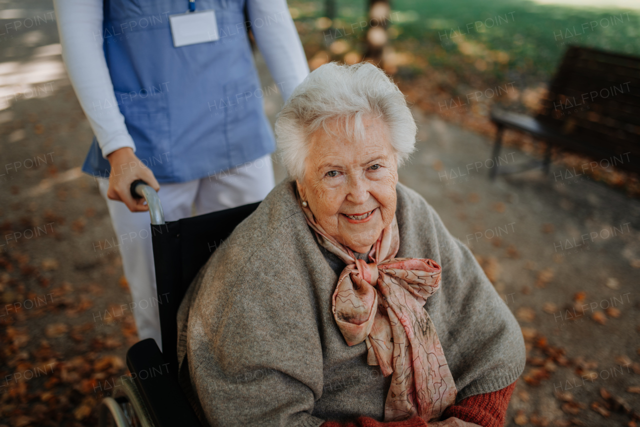 Disabled elderly woman in wheelchair on walk in park during warm autumn day. Young nurse pushing wheelchair, talking with senior woman.