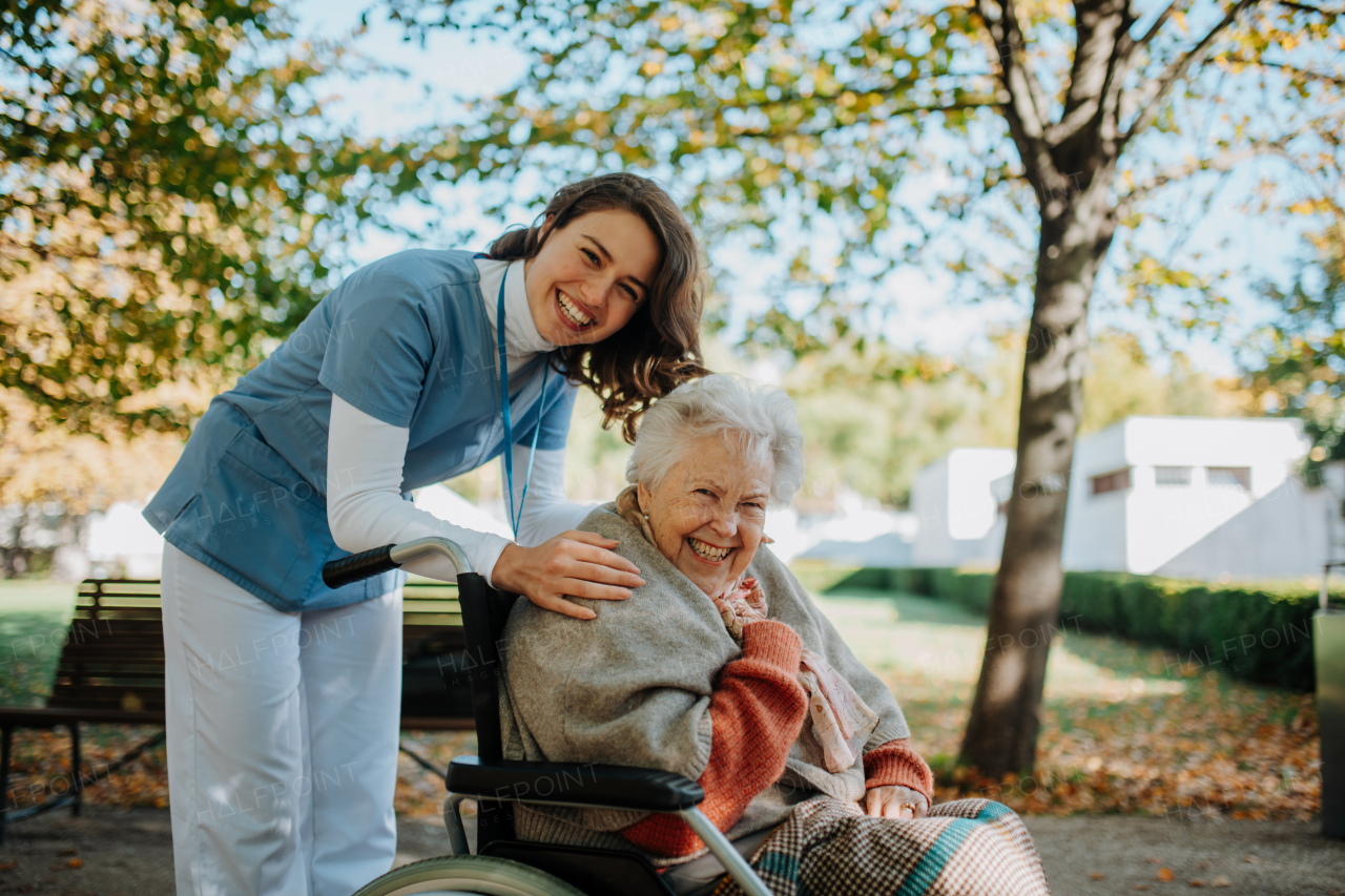 Disabled elderly woman in wheelchair on walk in park during warm autumn day. Young nurse pushing wheelchair, talking with senior woman.