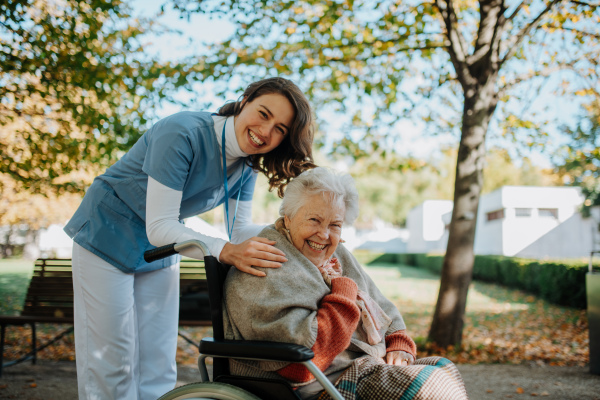 Disabled elderly woman in wheelchair on walk in park during warm autumn day. Young nurse pushing wheelchair, talking with senior woman.