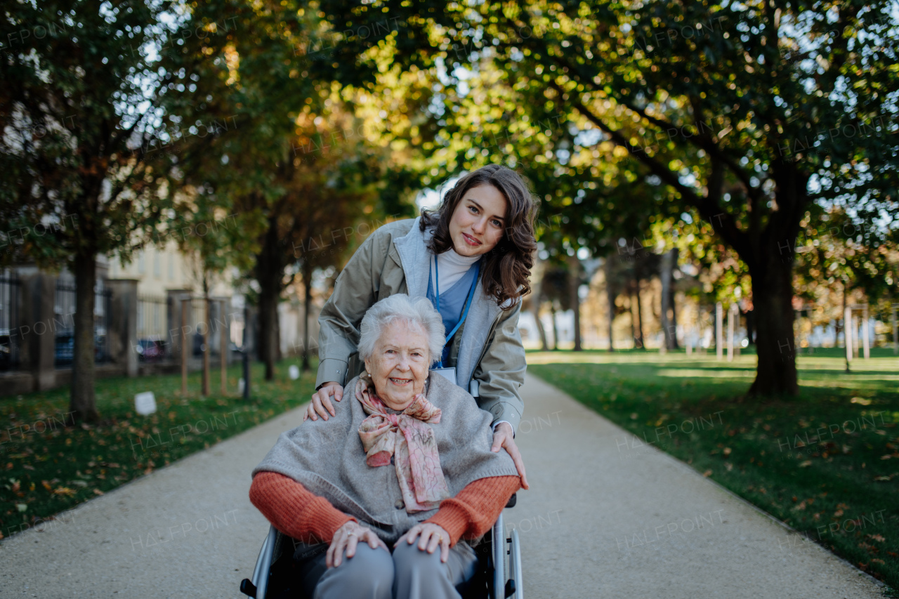 Disabled elderly woman in wheelchair on walk in park during warm autumn day. Young nurse pushing wheelchair, talking with senior woman.