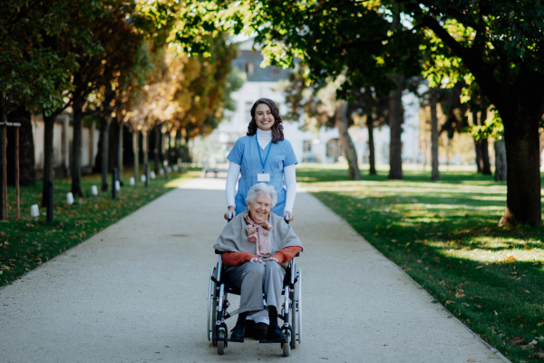 Disabled elderly woman in wheelchair on walk in park during warm autumn day. Young nurse pushing wheelchair, talking with senior woman.