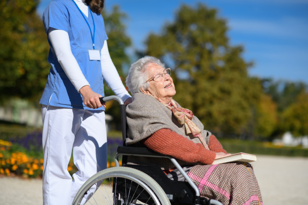 Disabled elderly woman in wheelchair on walk in park during warm autumn day. Young nurse pushing wheelchair, talking with senior woman.