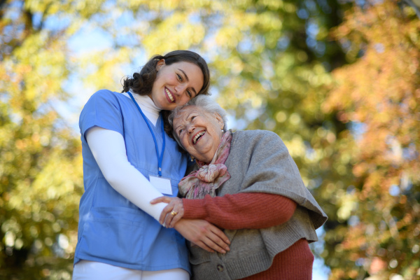 Portrait of nurse and elderly woman on walk in park during warm autumn day. Young caregiver spending time with senior patient.