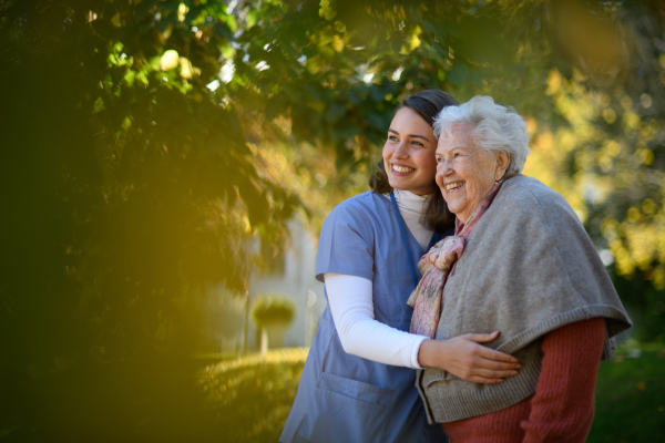 Portrait of nurse and elderly woman on walk in park during warm autumn day. Young caregiver spending time with senior patient.