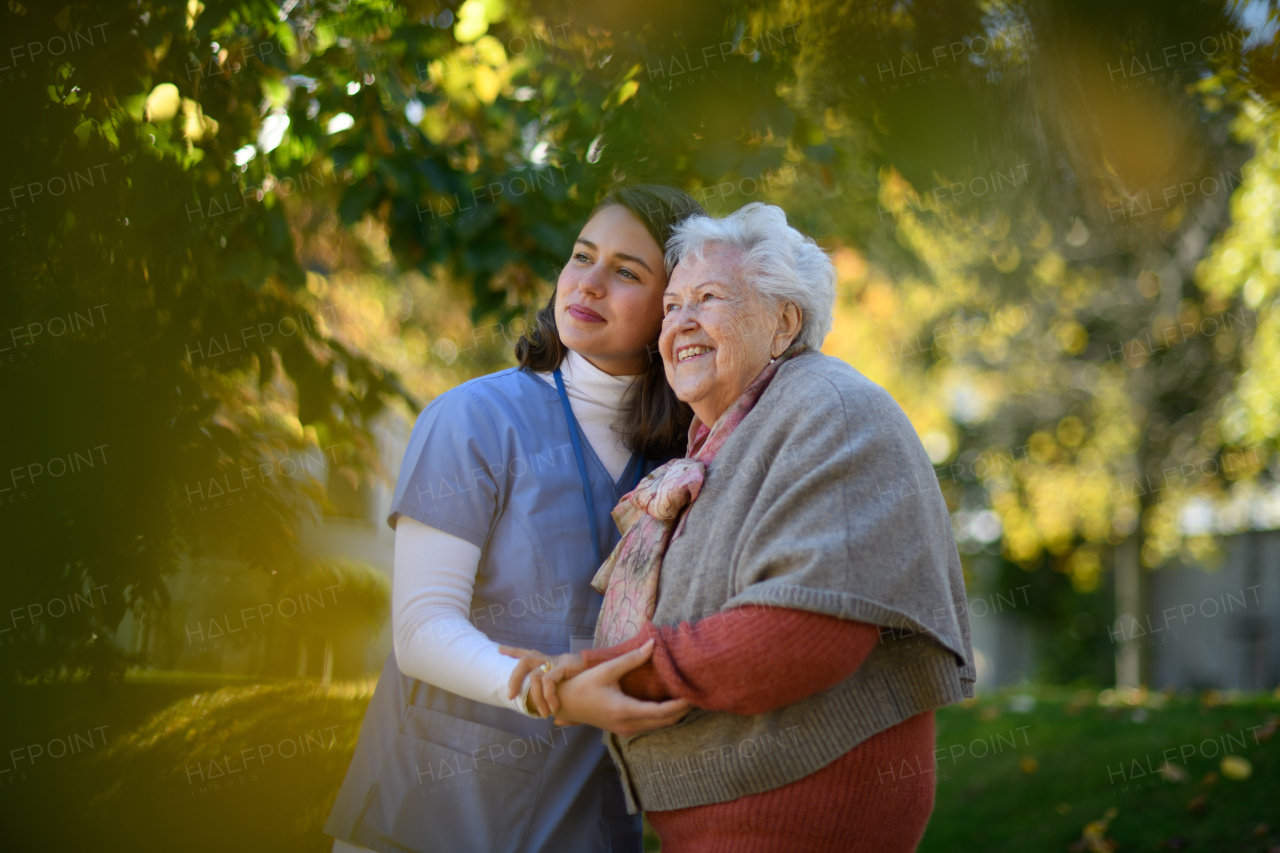 Portrait of nurse and elderly woman on walk in park during warm autumn day. Young caregiver spending time with senior patient.