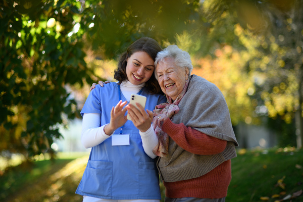 Portrait of nurse and elderly woman on walk in park during warm autumn day. Young caregiver looking through photos of senior patient's family on a phone