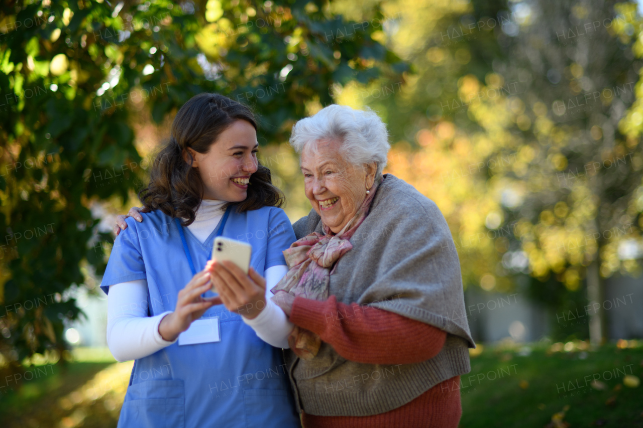 Portrait of nurse and elderly woman on walk in park during warm autumn day. Young caregiver looking through photos of senior patient's family on a phone