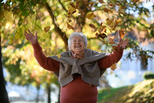 Portrait of elderly woman on walk in the park enjoying warm autumn day, throwing colorful leaves in the air.