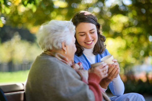 Elderly woman enjoying cup of coffee with her nurse in park during warm autumn day. Young caregiver spending time with senior woman.