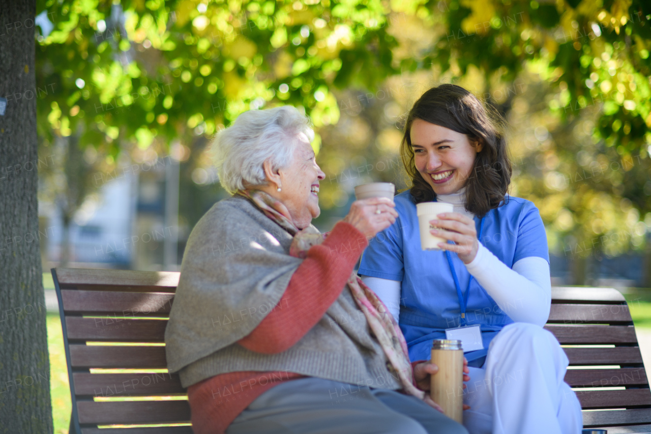 Elderly woman enjoying cup of coffee with her nurse in park during warm autumn day. Young caregiver spending time with senior woman.