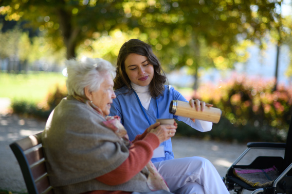 Elderly woman enjoying cup of coffee with her nurse in park during warm autumn day. Young caregiver spending time with senior woman.