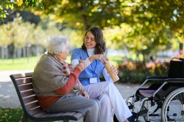 Elderly woman enjoying cup of coffee with her nurse in park during warm autumn day. Young caregiver spending time with senior woman.