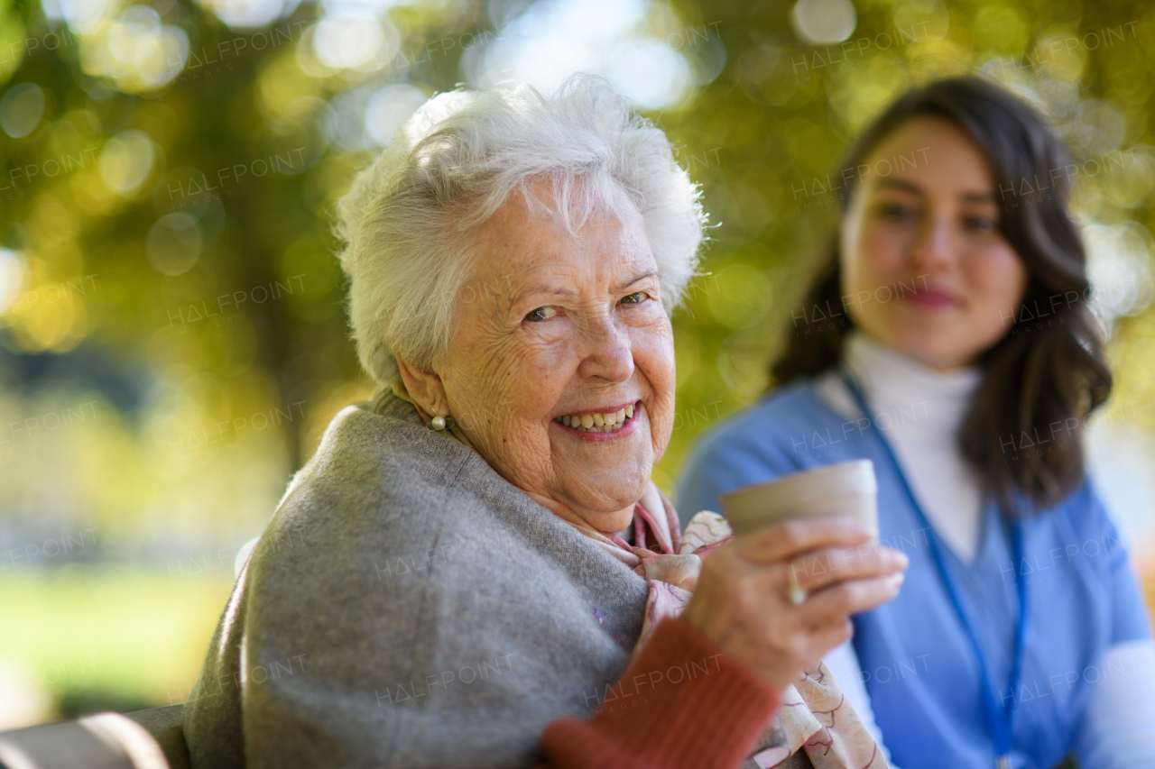 Elderly woman enjoying cup of coffee with her nurse in park during warm autumn day. Young caregiver spending time with senior woman.