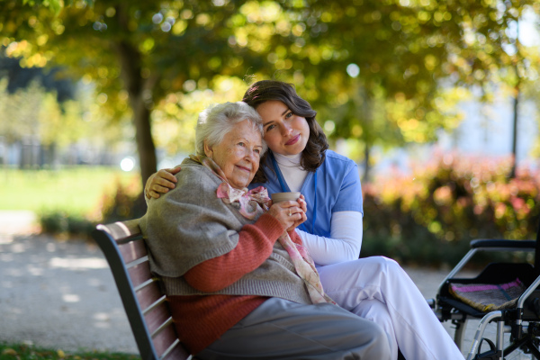 Elderly woman enjoying cup of coffee with her nurse in park during warm autumn day. Young caregiver spending time with senior woman.