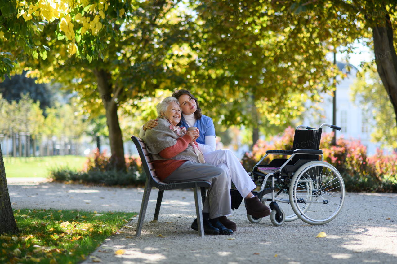 Elderly woman enjoying cup of coffee with her nurse in park during warm autumn day. Young caregiver spending time with senior woman.