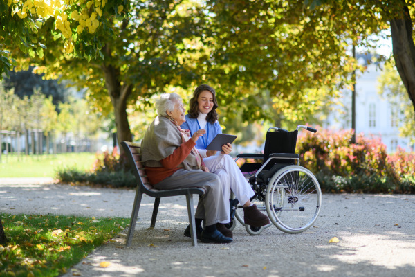 Portrait of nurse and elderly woman on walk in park during warm autumn day. Young caregiver looking through photos of senior patient's family on tablet