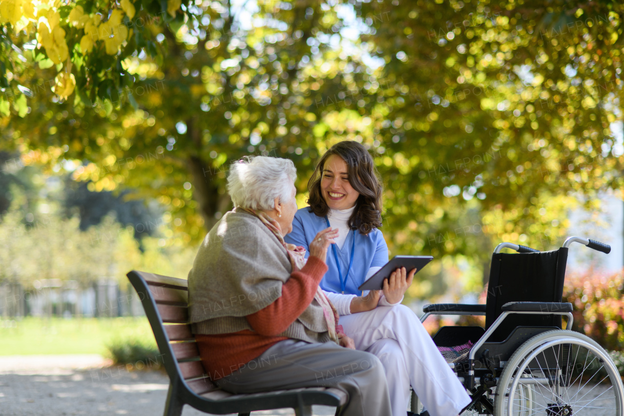 Portrait of nurse and elderly woman sitting on bench in park during warm autumn day. Young caregiver looking through photos of senior patient's family on tablet