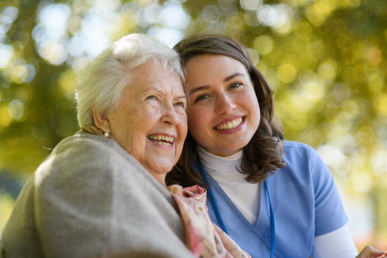 Portrait of nurse and elderly woman on walk in park during warm autumn day. Young caregiver spending time with senior patient.