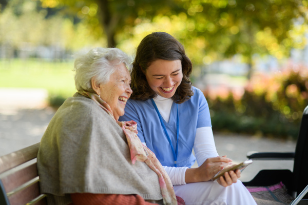 Portrait of nurse and elderly woman on walk in park during warm autumn day. Young caregiver looking through photos of senior patient's family on a phone