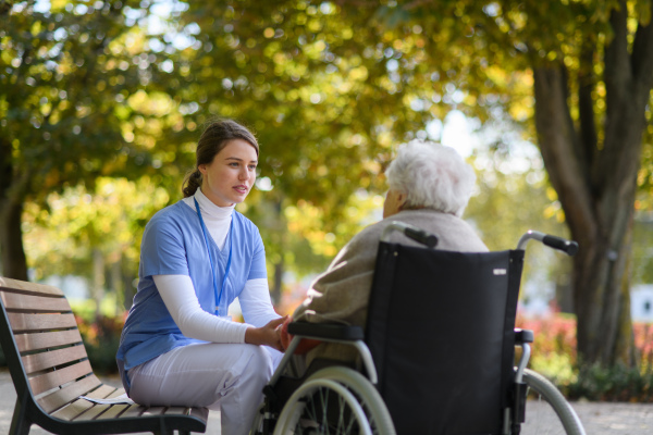 Disabled elderly woman in wheelchair holding hands with her nurse and talking about her problems.