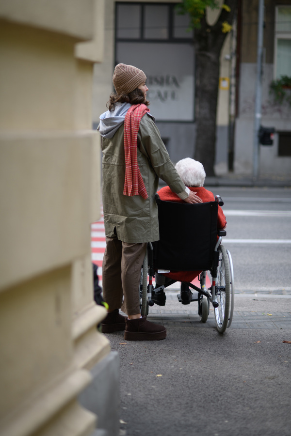 Granddaughter on autumn walk in the city with her grandmother, pushing her in wheelchair. Rear view.