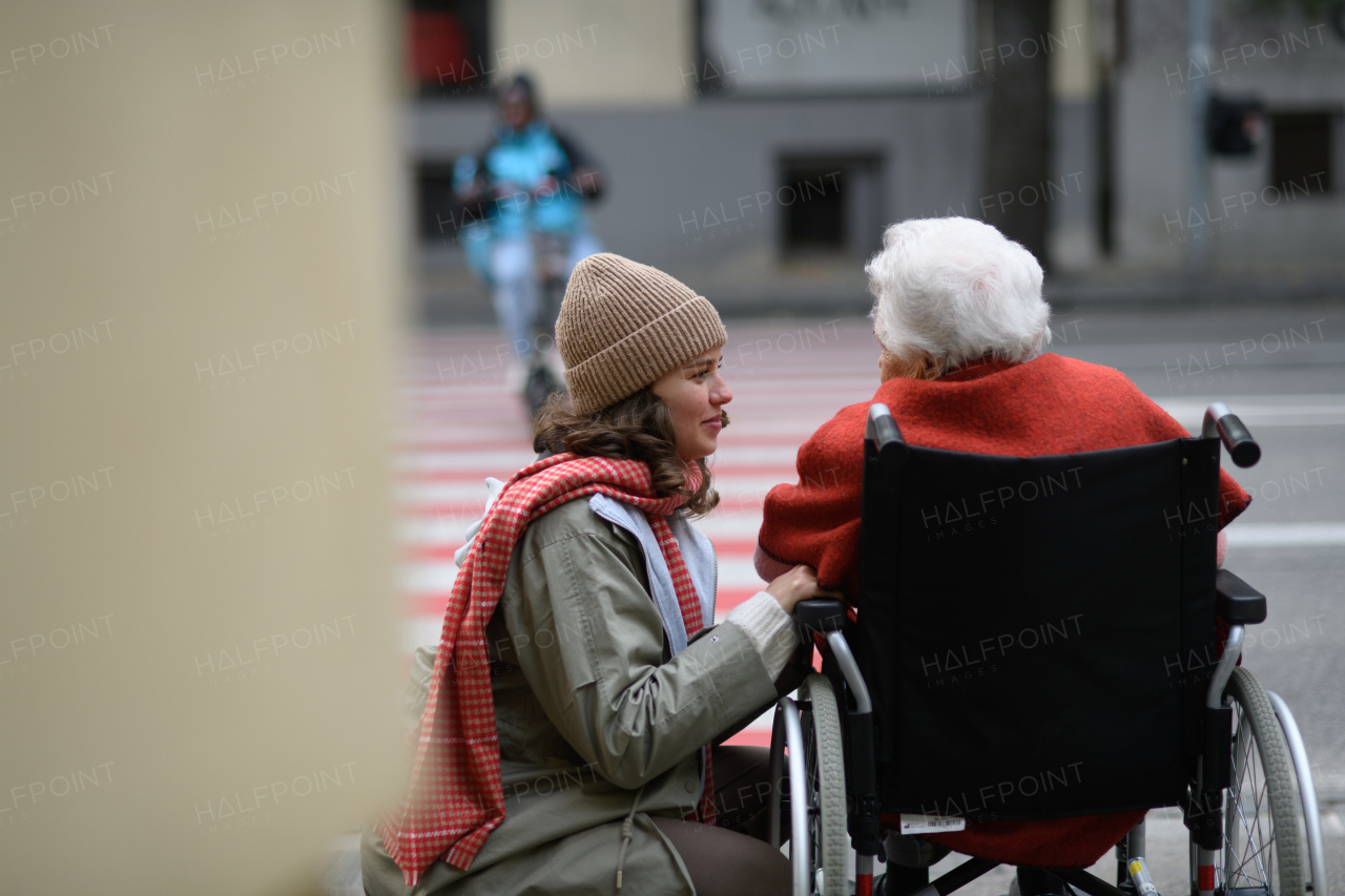 Granddaughter on autumn walk in the city with her grandmother, pushing her in wheelchair. Rear view.