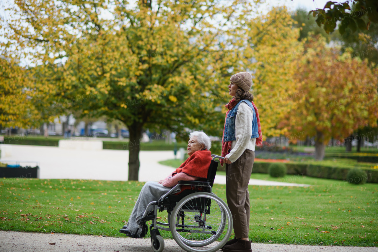 Granddaughter on an autumn walk in the park with her grandmother, pushing her in wheelchair.