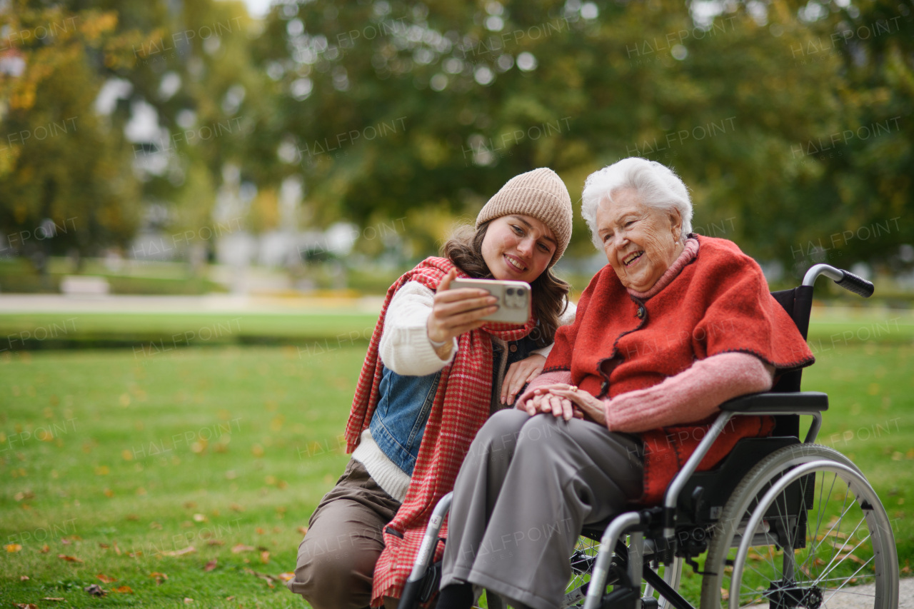 Granddaughter on an autumn walk in the park with her grandmother in a wheelchair, taking selfie with smartphone