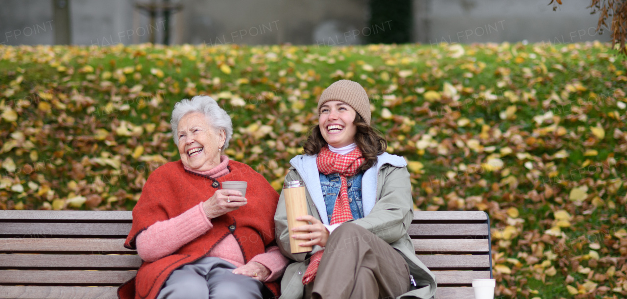 Portrait of granddaughter and grandmother sitting on bench in autumn park, drinking coffee, laughing together