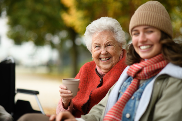 Portrait of granddaughter and grandmother sitting on bench in autumn park, drinking coffee and talking.