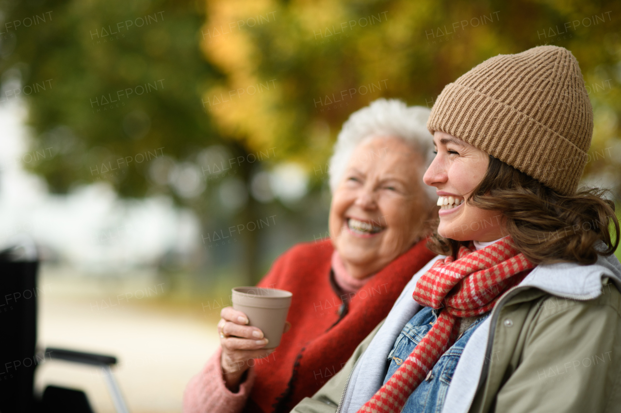 Portrait of granddaughter and grandmother sitting on bench in autumn park, drinking coffee and talking.