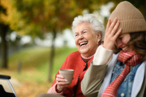 Portrait of granddaughter and grandmother sitting on bench in autumn park, drinking coffee, laughing together