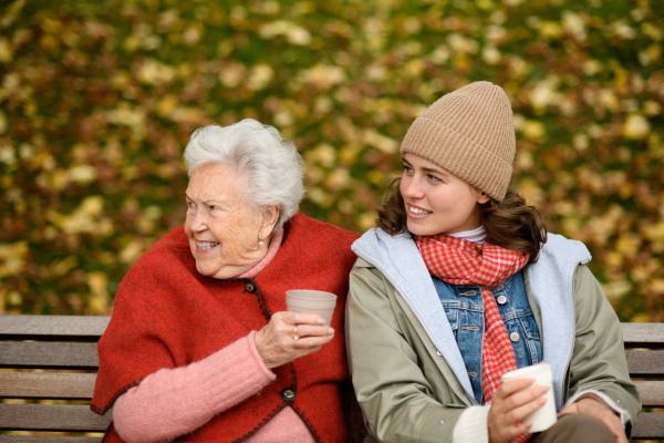 Portrait of granddaughter and grandmother sitting on bench in autumn park, drinking coffee, laughing together