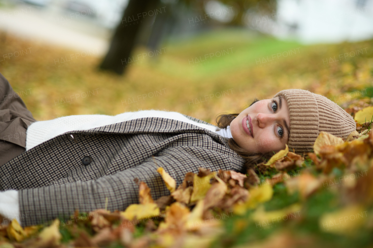 Portrait of beautiful woman lying in fallen autumn leaves on the lawn. She smiles and looking at camera.