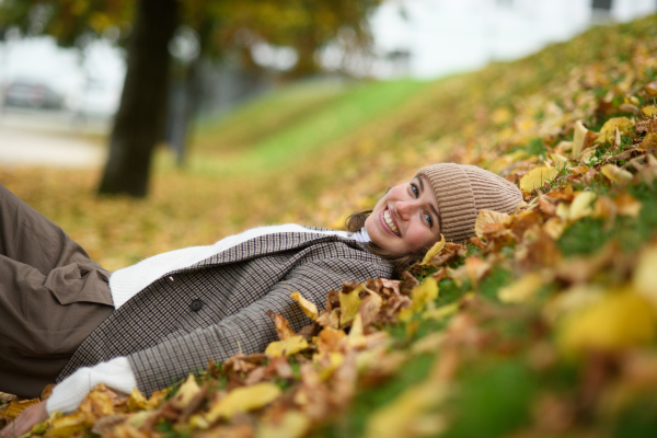 Portrait of beautiful woman lying in fallen autumn leaves on the lawn. She smiles and looking at camera.