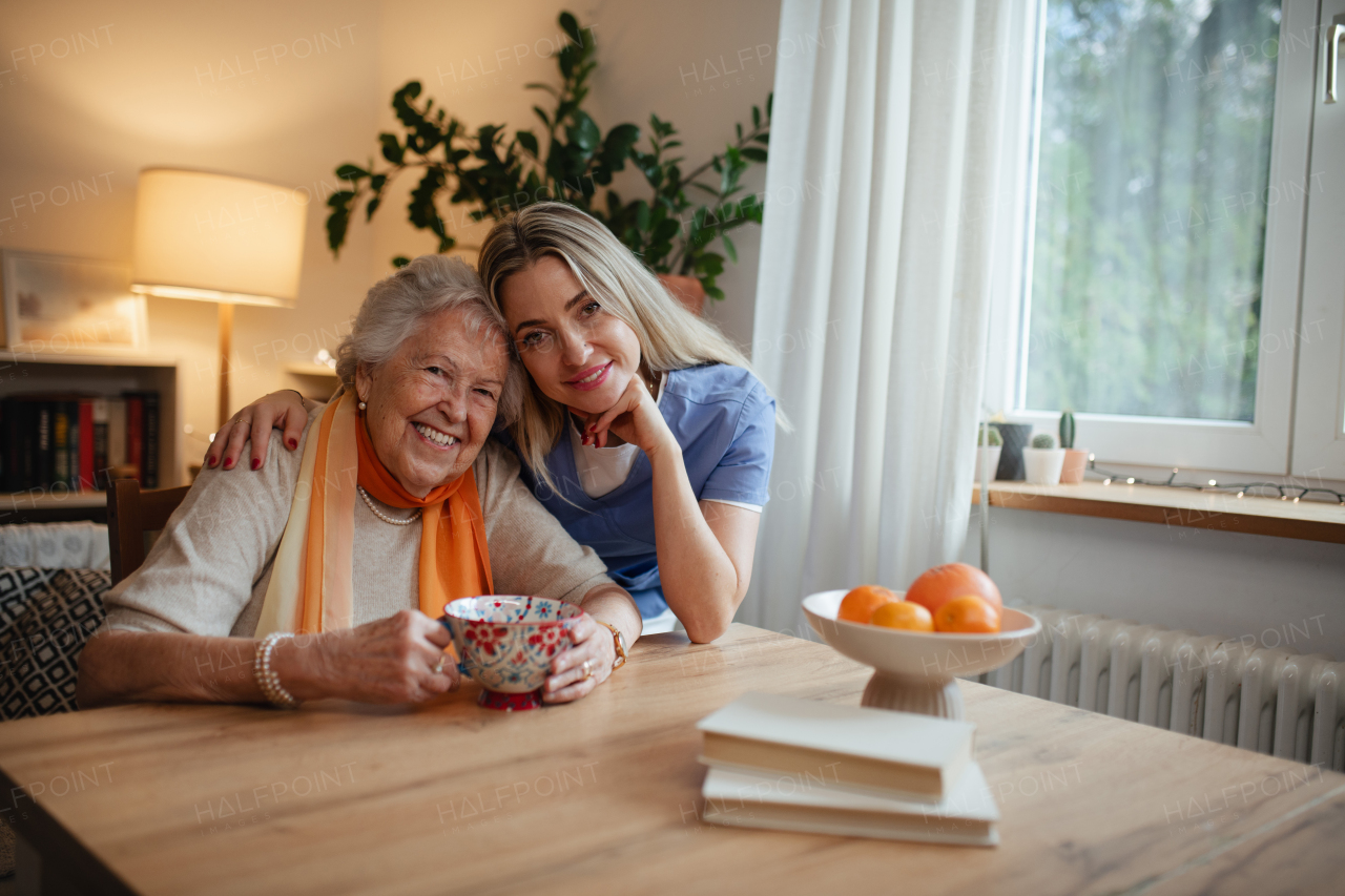 Caring healthcare worker visiting a senior patient at home. Lovely elderly woman sitting at table, looking at camera.