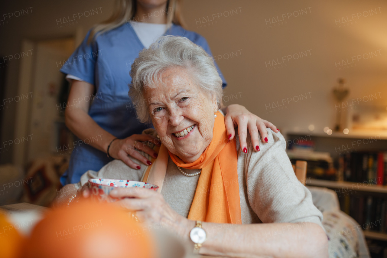 Caring healthcare worker visiting a senior patient at home. Lovely elderly woman sitting at table, looking at camera.