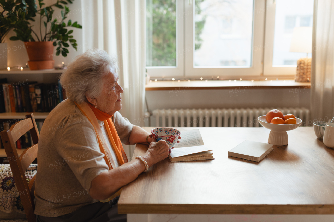 Side view of elderly lady reading a book, sitting at the kitchen table and her sipping tea.