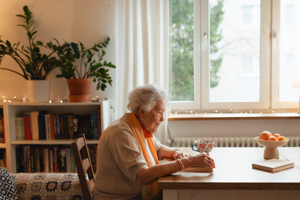 Side view of elderly lady reading a book, sitting at the kitchen table and her sipping tea.