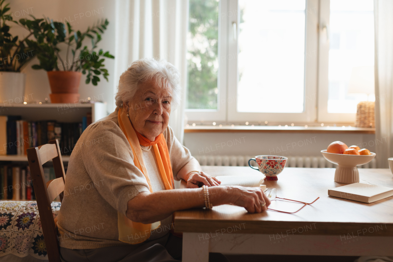 Side view of elderly lady reading a book, sitting at the kitchen table and her sipping tea.