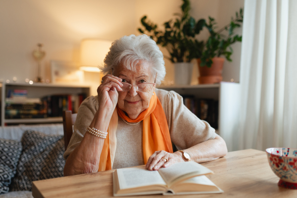 Side view of elderly lady reading a book, sitting at the kitchen table and her sipping tea.