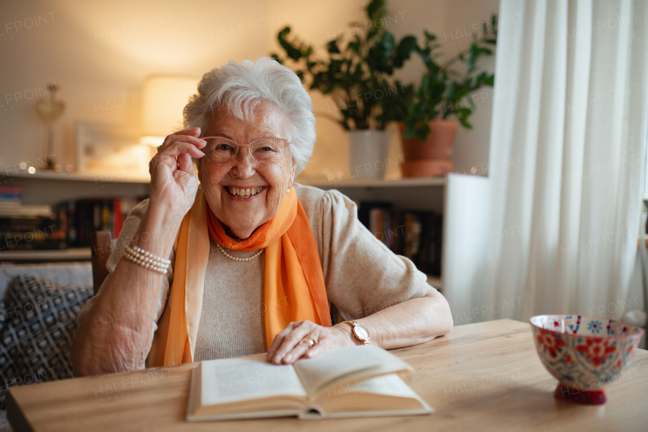 Lovely elderly lady reading book, sitting at the kitchen table and sipping tea.