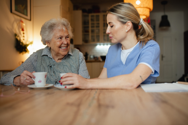 Lovely eldery woman serving tea to friendly caregiver during home visit. Home health care services.