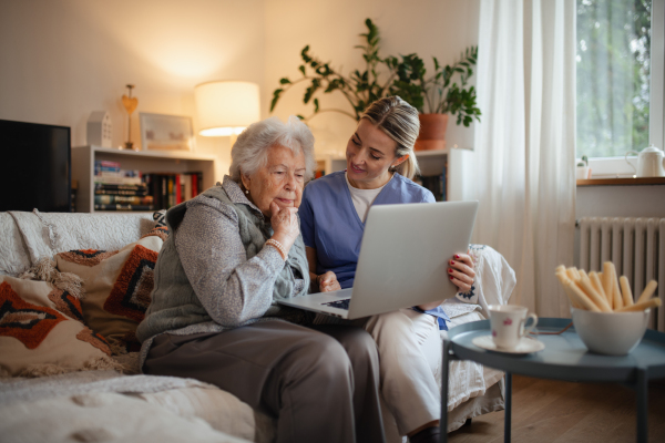 Social worker helping elderly lady with managing her finances and teaching her to use online banking on laptop.