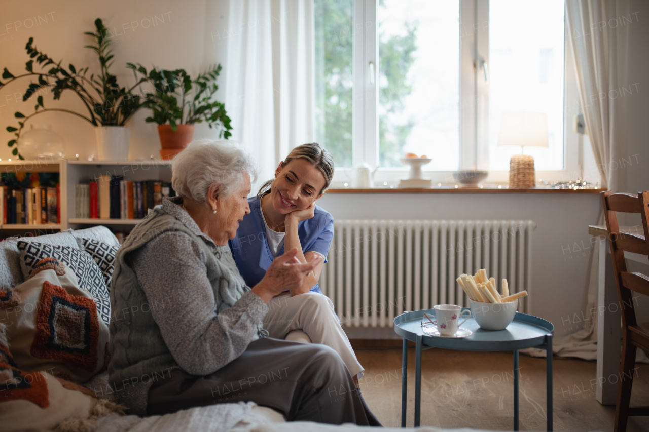 Caring healthcare worker visiting senior patient at home, talking with her in living room.