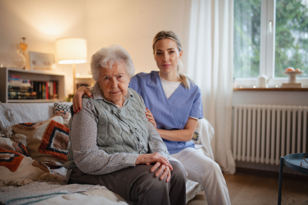 Caring healthcare worker visiting senior patient at home, sitting on sofa and looking at camera.