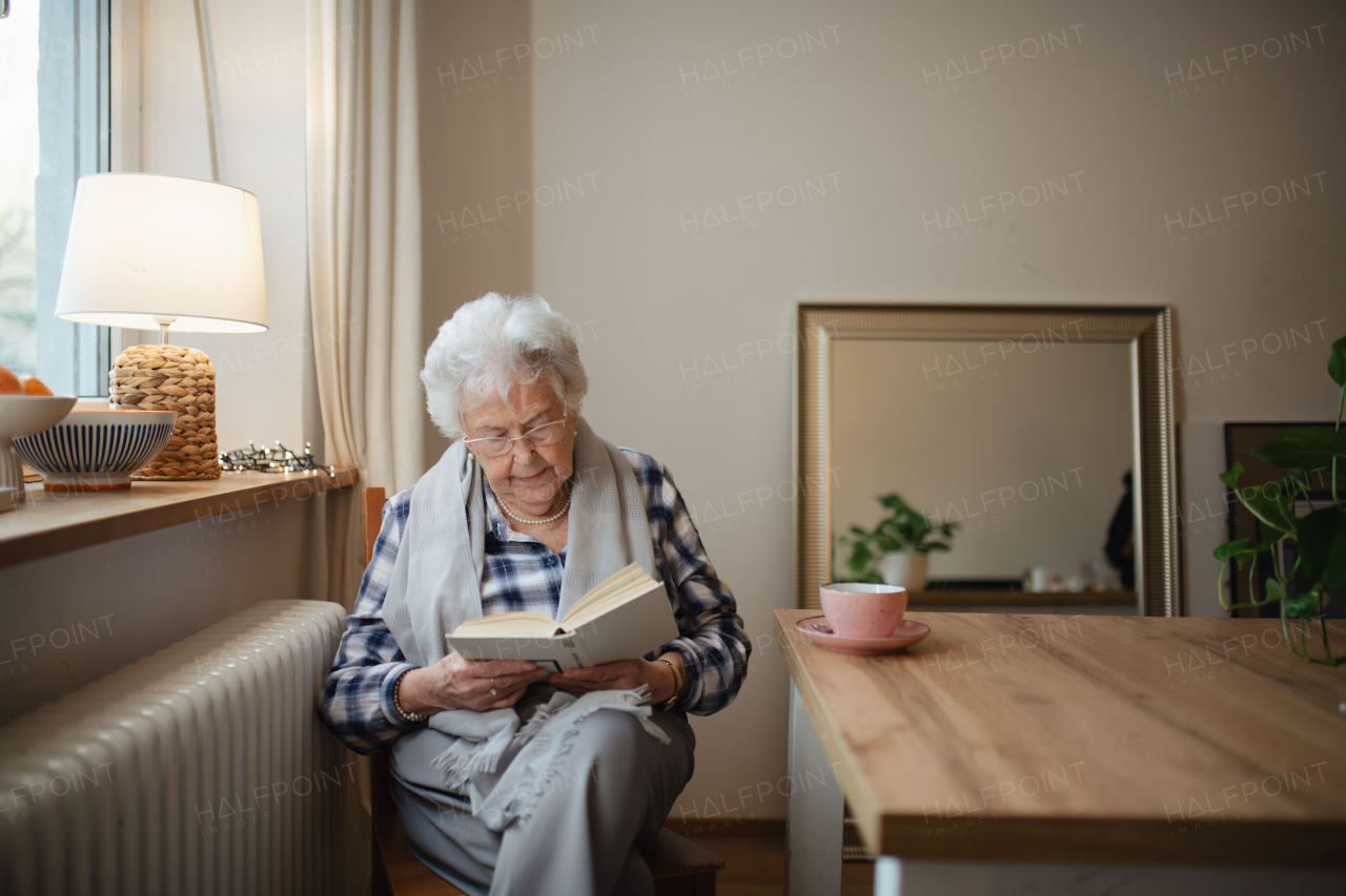 Lovely elderly lady reading book, sitting at the kitchen table and sipping tea.