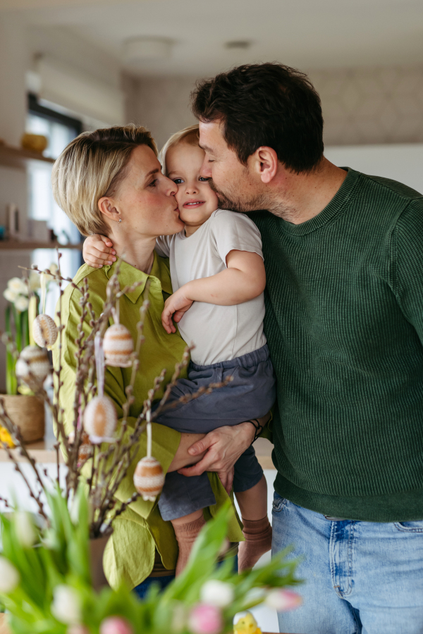 Parents standing next to pussy willows branches with easter eggs on them, kissing little boy on cheeks.