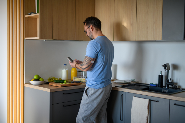 Diabetic man with CGM sensor standing in kitchen and checking his blood sugar level while making healthy breakfast.